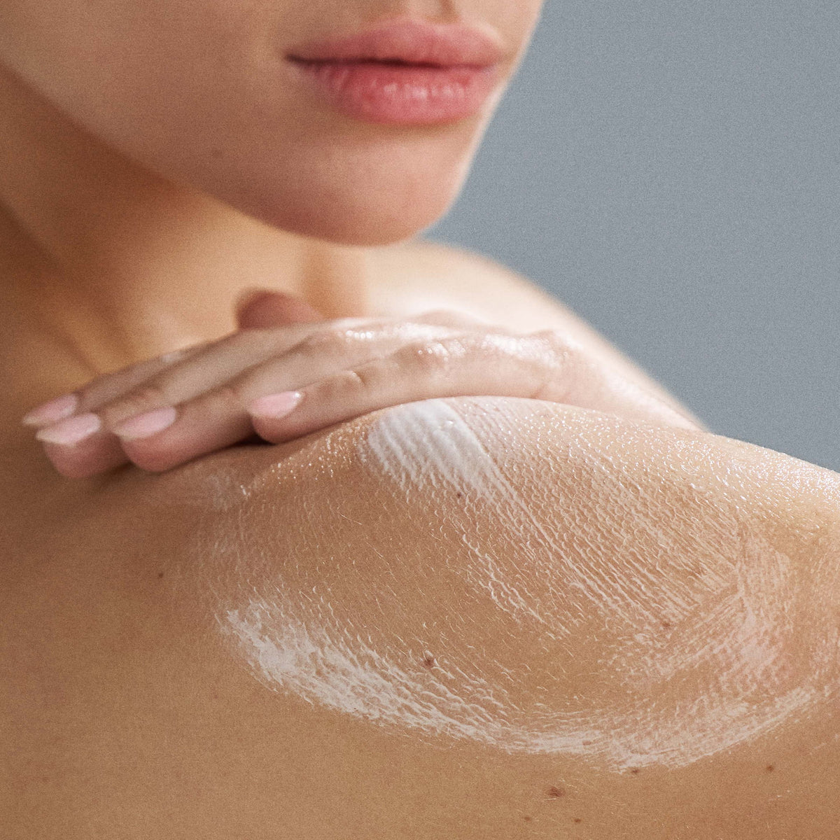 Close-up of a person applying body cream to their shoulder with a neutral background 