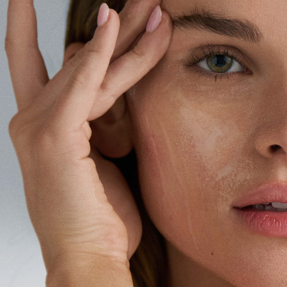 Close-up of a woman's face with one hand gently touching her cheek  that has a cleanser against a neutral background 